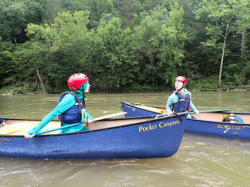 Two people wearing helmets and life jackets are paddling blue canoes on a river. The canoe in the foreground has the words "Pocket Canyon" written on its side. Lush green trees line the riverbank in the background. The water is calm, and the sky is overcast.